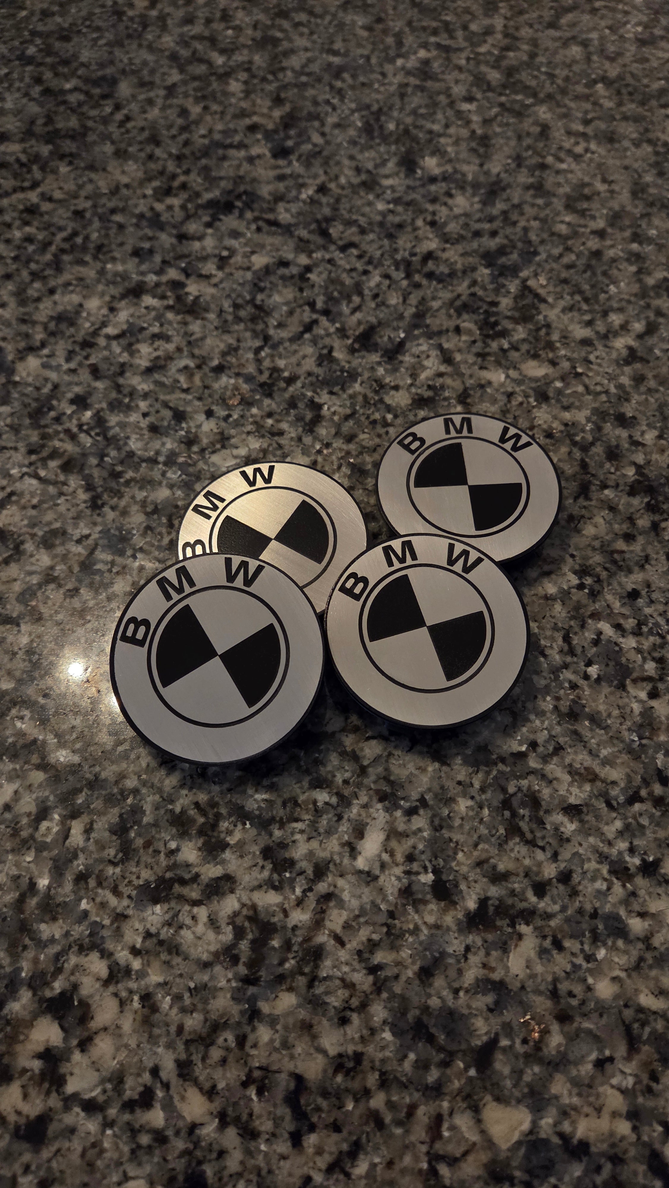 Four round BMW logo badges or emblems with black and white checkered centers and surrounding BMW lettering, placed on a dark speckled granite surface.